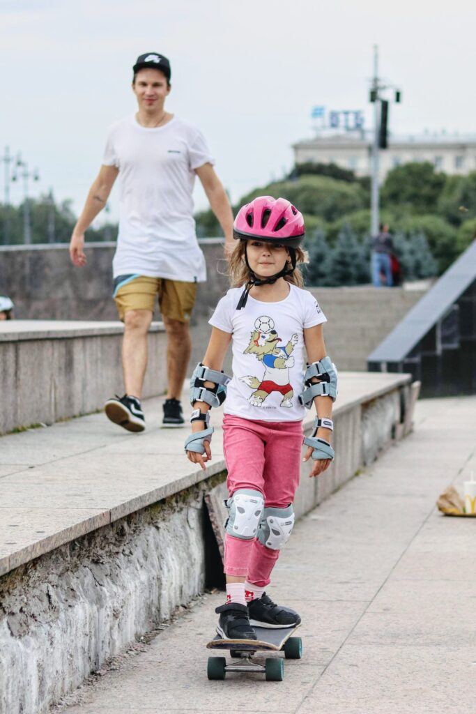 Young student in a pink helmet riding a skateboard during a One2Flip lesson while Vladimir watches